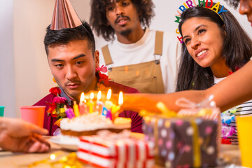 Multi-ethnic group of friends at a birthday party on the sofa at home with a cake and gifts, lighting the candles