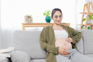 Obraz premium pregnant asian people sitting on sofa, she use her hand touch on her abdomen, she feeling love and care her daughter, pregnancy health care promotion
