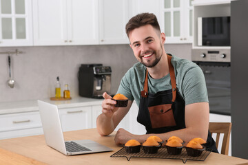 Happy man holding muffin near laptop at table in kitchen. Time for hobby