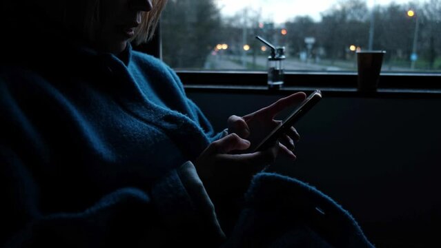 A woman is chatting on the phone in a dark room near the window. Evening time of day. Loneliness, search for acquaintances.