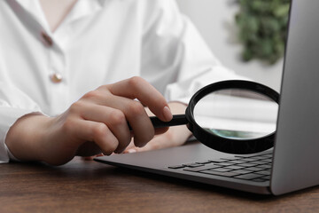 Woman holding magnifier near laptop at wooden table indoors, closeup. Online searching concept