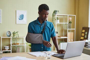Portrait of black young man with arm sling working at standing desk and using laptop in office