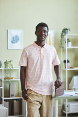 Vertical portrait of young black man smiling at camera standing by desk in pastel colored office