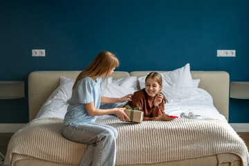 Happy surprised adolescent teen girl receiving birthday present from mother while resting on bed. Loving mom giving wrapped gift box to excited teenage daughter, congratulating child at home