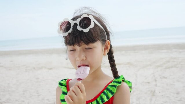 Little Asian child girl in swimsuit eating ice cream during playing with family on tropical beach in sunny day. Happy children kid enjoy and fun outdoor activity lifestyle on summer holiday vacation