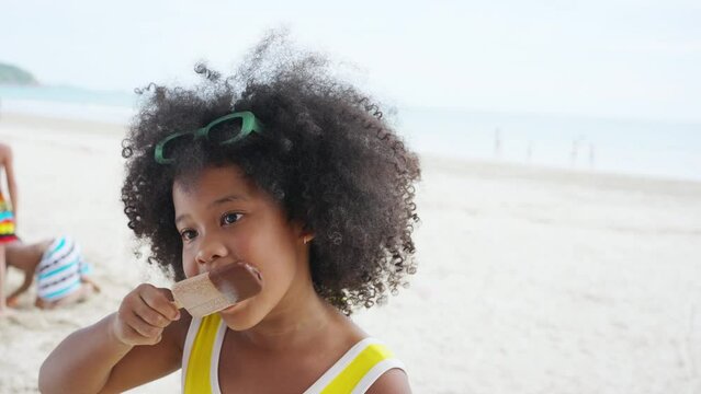 Little African child girl in swimsuit eating ice cream during playing with family on tropical beach in sunny day. Happy children kid enjoy and fun outdoor activity lifestyle on summer holiday vacation