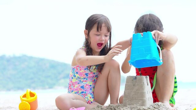 Little cute child girl friends in swimsuit playing beach toy and build a sand castle together at tropical beach. Children kids enjoy and fun outdoor activity lifestyle travel ocean on summer vacation.