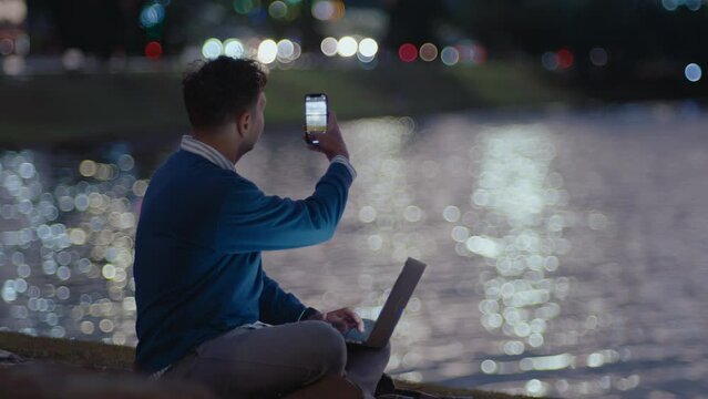Young man sitting in city park by the river, working online on laptop, taking photos of night city with smartphone in the evening. Bokeh car lights and reflecting water surface in the background