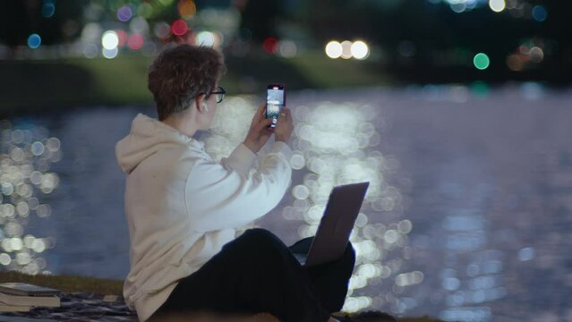Young woman sitting in the park by the river, doing college homework on laptop, taking pictures of the city with smartphone in the evening. Bokeh lights on reflecting water surface in the background