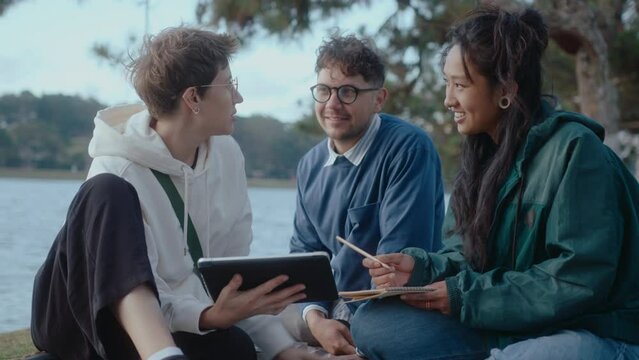 Company of diverse college students sitting in the park, using a digital tablet, discussing homework or working on a group project together. Medium full shot