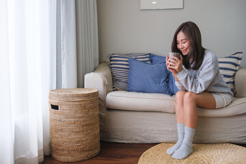 Portrait image of a beautiful asian woman with closed eyes drinking hot coffee and relaxing on a sofa at home