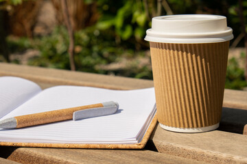 Eco recycling paper cup with coffee or tea on kraft paper with empty paper notebook on wooden bench. Concept of study work outdoors. Take away coffee to go. Copy space for text. Disposable Cardboard