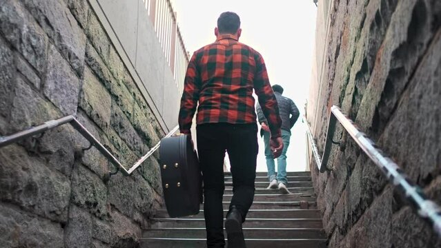 Musician is walking up the stairs holding a guitar case and has people walking in front of him.