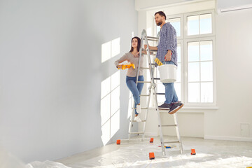 Happy young couple painting the wall of their new home holding paint rollers and standing on the...
