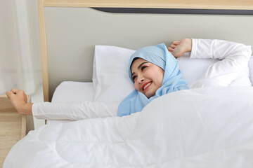 Fototapeta na wymiar Beautiful asian woman wearing white muslim sleepwear lying on bed, stretching her arms after getting up in the morning at sunrise. Cute young girl with hijab wake up and relaxing, closing her eyes
