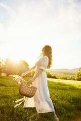 a woman in a light dress in a chamomile field against the background of a sunset stands with her...