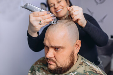 Fototapeta premium Adult man with beard in camouflage T-shirt receiving bald shave service before going to army. Girl gets rid of her hair with electric razor.