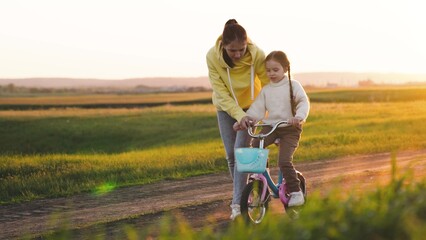 mother teaches child ride two-wheeled bicycle sunset. happy family. chidhood dream. girl with mom ride bike outdoors. happy little girl daughter rides rural road sunset. concept family sports road - obrazy, fototapety, plakaty
