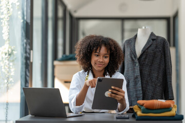 African fashion designer or dressmaker American uses ideas and presents work on designing new collections with fabric samples and sketches on a tablet working in the studio.