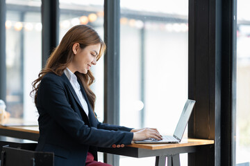 Confident young asian businesswoman sitting on chair in front of counter recording plan online business financial goals Data entry on laptop of successful woman