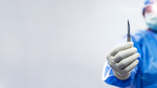Male doctor or surgeon in blue gown holding surgical scalpel or medical knife with blur white wall background.Surgical blade for cosmetic or orthopedic surgery with space.Man wearing loupe with light.