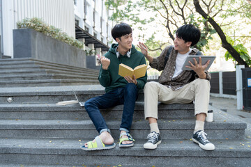 Fototapeta premium Two Asian male students sitting on the stairs of the university enjoy chatting after school using smartphones and tablets to find information together.