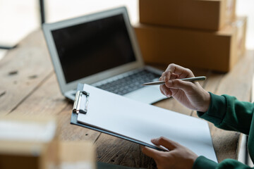 Fototapeta premium starting a small business SME, Asian businesswoman or e-commerce store owner using laptop and chart to check orders, online ordering, shipping tracking number to prepare packages in office.