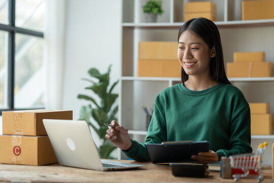 starting a small business SME, Asian businesswoman or e-commerce store owner using laptop and chart to check orders, online ordering, shipping tracking number to prepare packages in office.