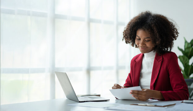 Project revenue and profit accounting African american businesswoman working with laptop financial charts and graphs checking and reading data details in financial paper documents at desk.