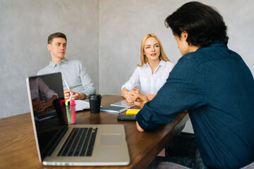 Group of three young businesspeople talking sitting at desk with laptop in light modern office. Friendly multi-ethnic business team discussing business issues at meeting. Concept of corporate meeting