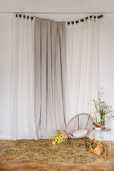 Сhair, table with flowers, wicker basket with apples on dry plant, straw, hay. Home decor for photo sessions. Arch, wall with linen curtains. Rural decoration. Easter zone with rustic spring design. © Serhii