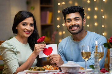 Happy smiling young boyfriend giving rose greeting to girlfriend by looking at camera during candle light dinner at home - concept of valentines day, relationship and romantic couple.
