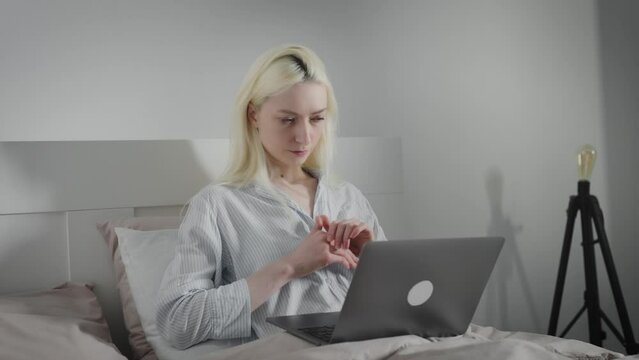 A blonde middle-aged woman uses a laptop lying in bed, typing on a computer keyboard