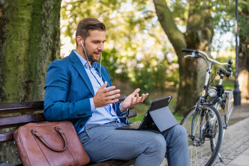 Fototapeta premium Caucasian male entrepreneur freelancer sitting outdoors in city park a bench talking online a video call using laptop or tablet. Business man office worker working on urban street background 