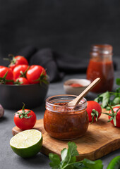 Sauce paste harissa with pepper and tomato in a jar on a dark background with fresh vegetables and herb close up. Adjika. Traditional italian, Georgian and Arabic cuisine.