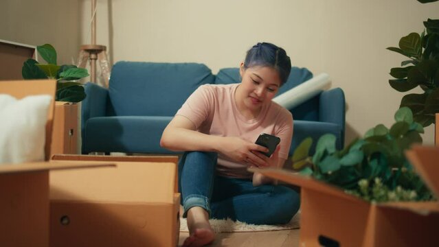 Young asian woman holding mobile phone in hand playing social media. Beautiful girl looking at smartphone chatting with friends and family in house moving day. Female using cellphone in living room.