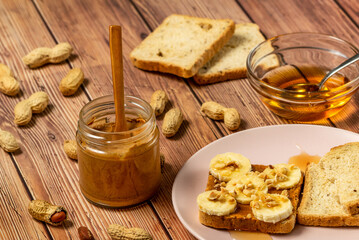 Peanut butter or peanut paste in an open jar and spread on a toast sandwich with banana and honey and peanuts in shells scattered on the table