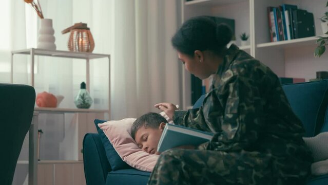 Loving mother in military uniform finishes reading book to son before sleep