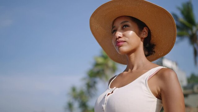 Black woman in straw hat looking at ocean at luxury tropical resort. Multiracial female relaxes on vacation to exotic island. Pretty bipoc girl enjoys sun on beach with palm trees.