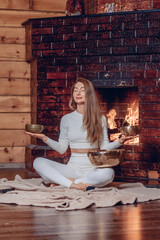 A wonderful woman meditates by a warm cozy fireplace sitting on a soft blanket with her eyes closed and holding several metal bowls for balance.