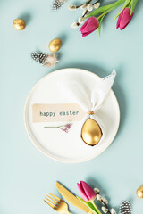 Stylish Easter flat lay with golden egg in easter bunny napkin on white plate, golden quail eggs, feathers, cutlery and spring flowers on blue background. Minimalist modern Easter background