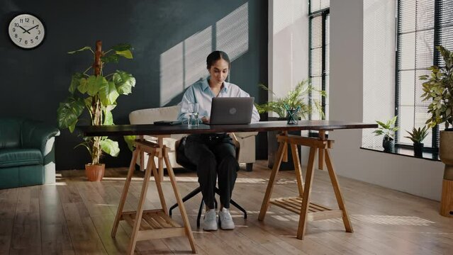 Full-length confident multi-ethnic woman, freelance entrepreneur sitting at dest, working on laptop