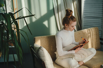 Satisfied ginger woman wearing white sweater sitting on cough writing in organizer, making to do list. Girl facial treatment. Creative idea. Health care. People