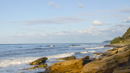 
Rocky seashore, waves crawl on huge boulders. Blue sky with clouds on the horizon.