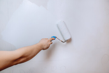 A man's hand holding a paint roller and priming the wall in white close-up. The concept of renovation in the apartment.