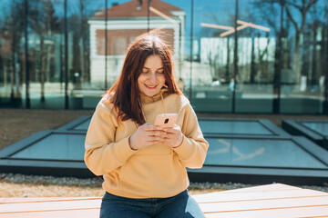 Beautiful woman in casual clothing using her smart phone while sitting on the bench on city street....