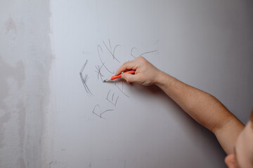 A man's hand writes obscene expressions in a foreign language with a graphite pencil on an empty white wall.