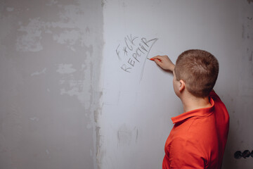 A young man standing with his back writes abusive words on an empty white wall with a graphite...