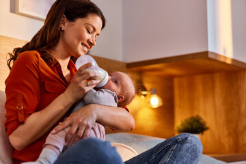 Sleepy cute infant child girl drinking milk from the bottle in mother's arms at home.