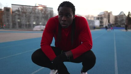 concentration on exercise. A young attractive African man in a red t-shirt rests squats on a running track. Initial body posture and sports discipline